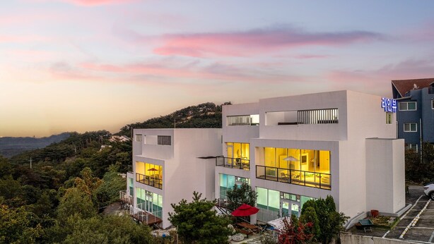 Un edificio moderno de varios pisos con balcones y ventanas amplias, rodeado de vegetación y colinas.