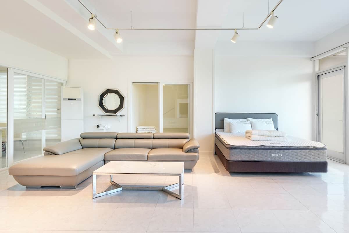 A modern bedroom with a sectional sofa, a bed with a mattress labeled 'BEREX', a small table, and a large window offering a view.