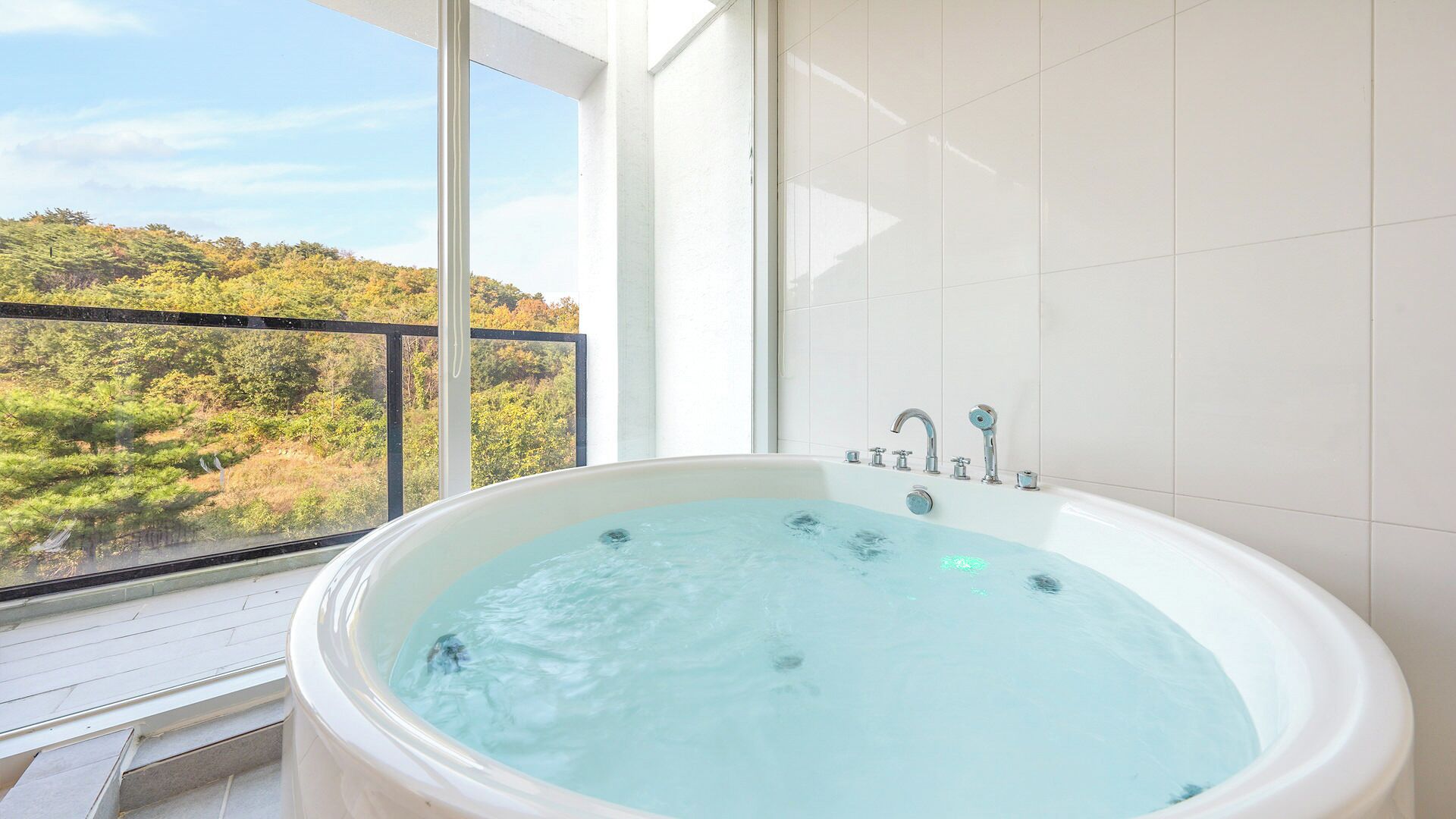 Un baño con una bañera grande, una ventana con vistas a un bosque y paredes de azulejos blancos.