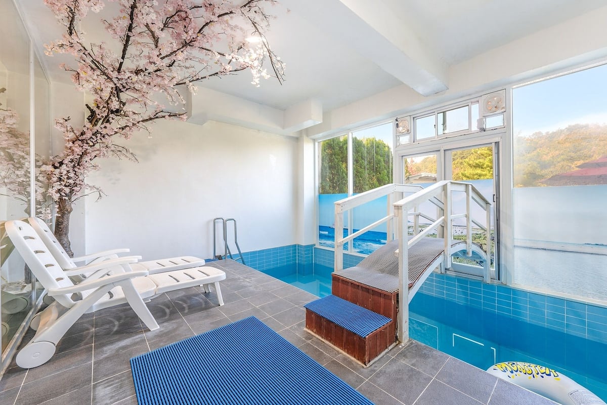 An indoor swimming pool with a view of a lake, a cherry blossom tree, and a wooden deck with lounge chairs.