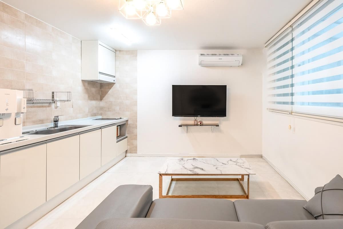 A compact living space with a kitchenette, a flat-screen TV mounted on the wall, and a window with blinds.
