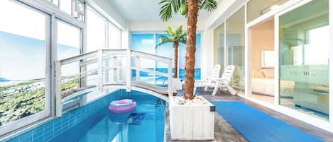 En indendørs swimmingpool med havudsigt, en palme og liggestole.