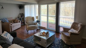 A spacious living room with a wooden floor, a beige armchair, a sofa, and a coffee table with magazines and a wooden sculpture. - Lovely 3BR/2BA Lakehouse Retreat in Gated Dubois, PA—Near Beach, Woods & Privacy (DuBois)
