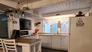 A compact kitchen with white cabinetry, a microwave, a stove, and a refrigerator. There are hanging lights and a window with a view of trees. - Lovely 3BR/2BA Lakehouse Retreat in Gated Dubois, PA—Near Beach, Woods & Privacy (DuBois)