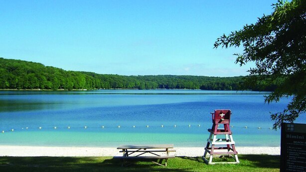 A lakeside view with a picnic table, a lifeguard chair, and a signboard. - Lovely 3BR/2BA Lakehouse Retreat in Gated Dubois, PA—Near Beach, Woods & Privacy (DuBois)