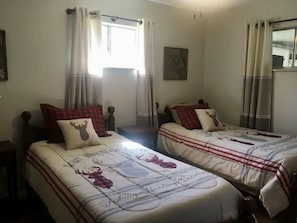 Una stanza con due letti, ciascuno con una coperta a quadri bianchi e rossi e un cuscino raffigurante una testa di cervo. Si vede una finestra con tende leggere, un quadro appeso al muro e uno specchio sopra il letto a destra.