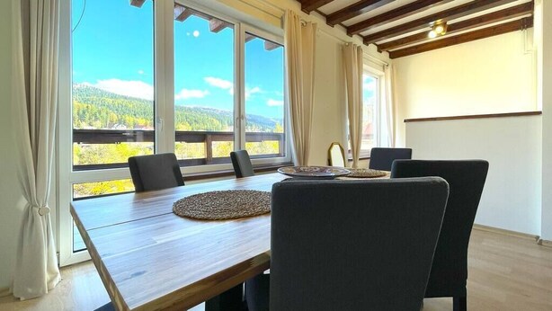 A dining area with a wooden table and chairs, large windows with a view of a forest and mountains, and a simple white wall with a light fixture. - Mountain Escape Chalet - BBQ and Mountain View (Bușteni)
