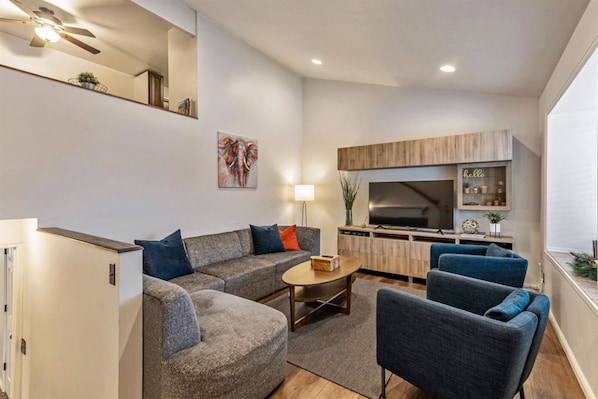A modern living room with a sofa, armchairs, and a coffee table. There is a TV, a cabinet, and a plant on the wall.