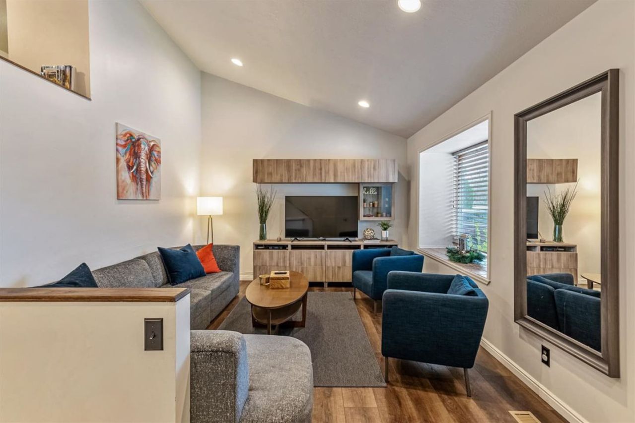 A modern living room with a sofa, armchairs, and a coffee table. There is a kitchen area with a TV and cabinets.