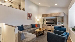Een moderne woonkamer met een bank, fauteuils en een salontafel. Er is een televisie, een kast en een plant aan de muur.