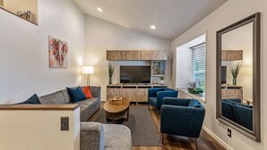 Una sala de estar moderna con un sofá, sillones y una mesa de centro. Hay una zona de cocina con un televisor y armarios.