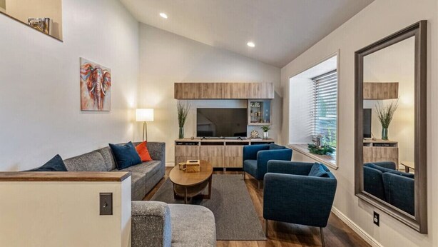Een moderne woonkamer met een bank, fauteuils en een salontafel. Er is een keukenhoek met een televisie en kasten.