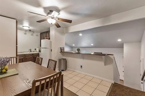 A dining area with a wooden table and chairs, a ceiling fan, recessed lighting, and a kitchen in the background.