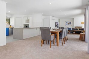 Une cuisine moderne avec des meubles blancs, un coin repas avec une table en bois et des chaises grises, et un salon avec un canapé et un tableau au mur.