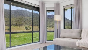 Une chambre moderne avec un canapé, une table basse en verre et de grandes fenêtres donnant sur un paysage verdoyant et des collines.
