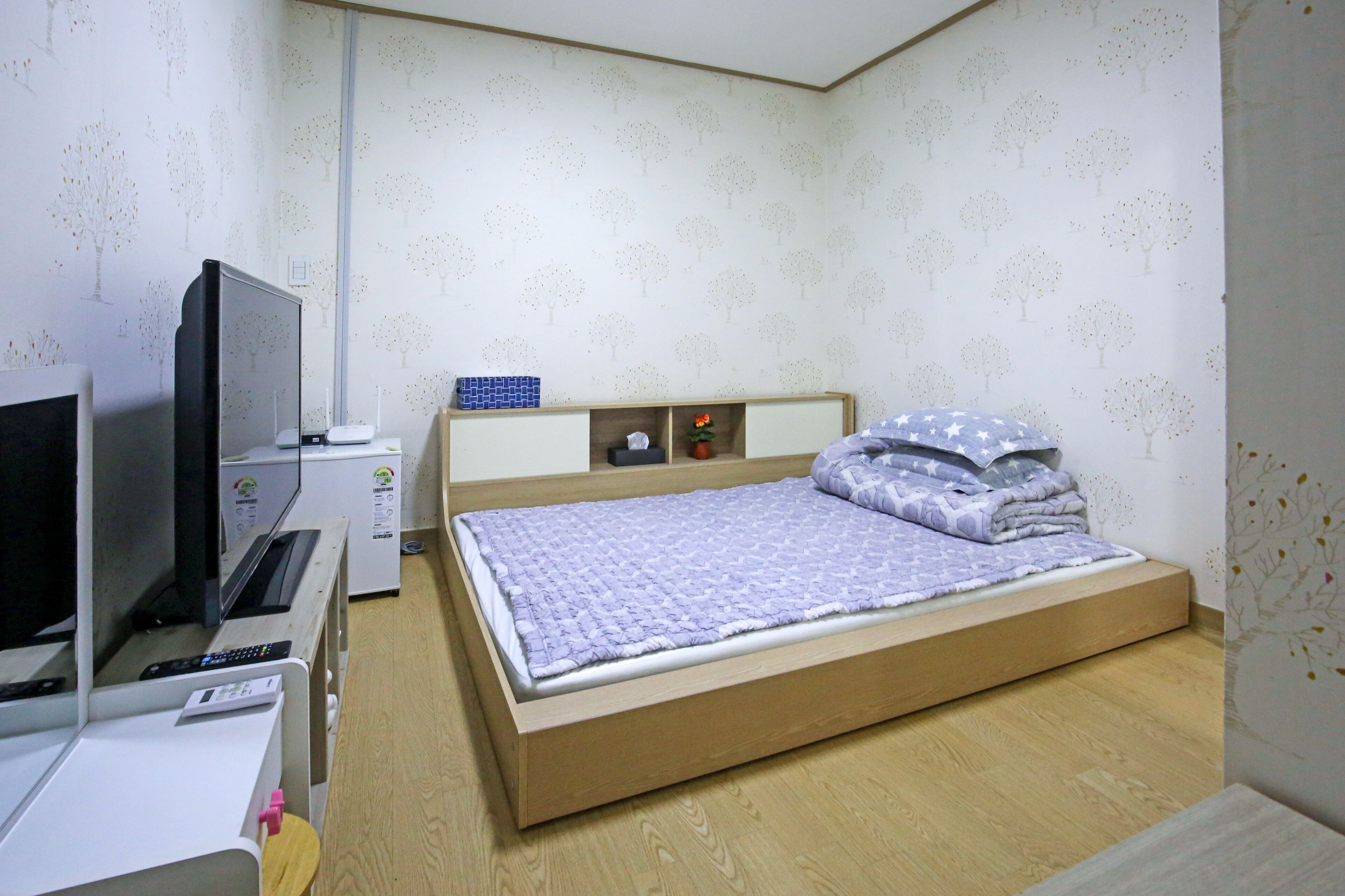 Un dormitorio con cama, televisor, escritorio con computadora y un estante pequeño con objetos decorativos.