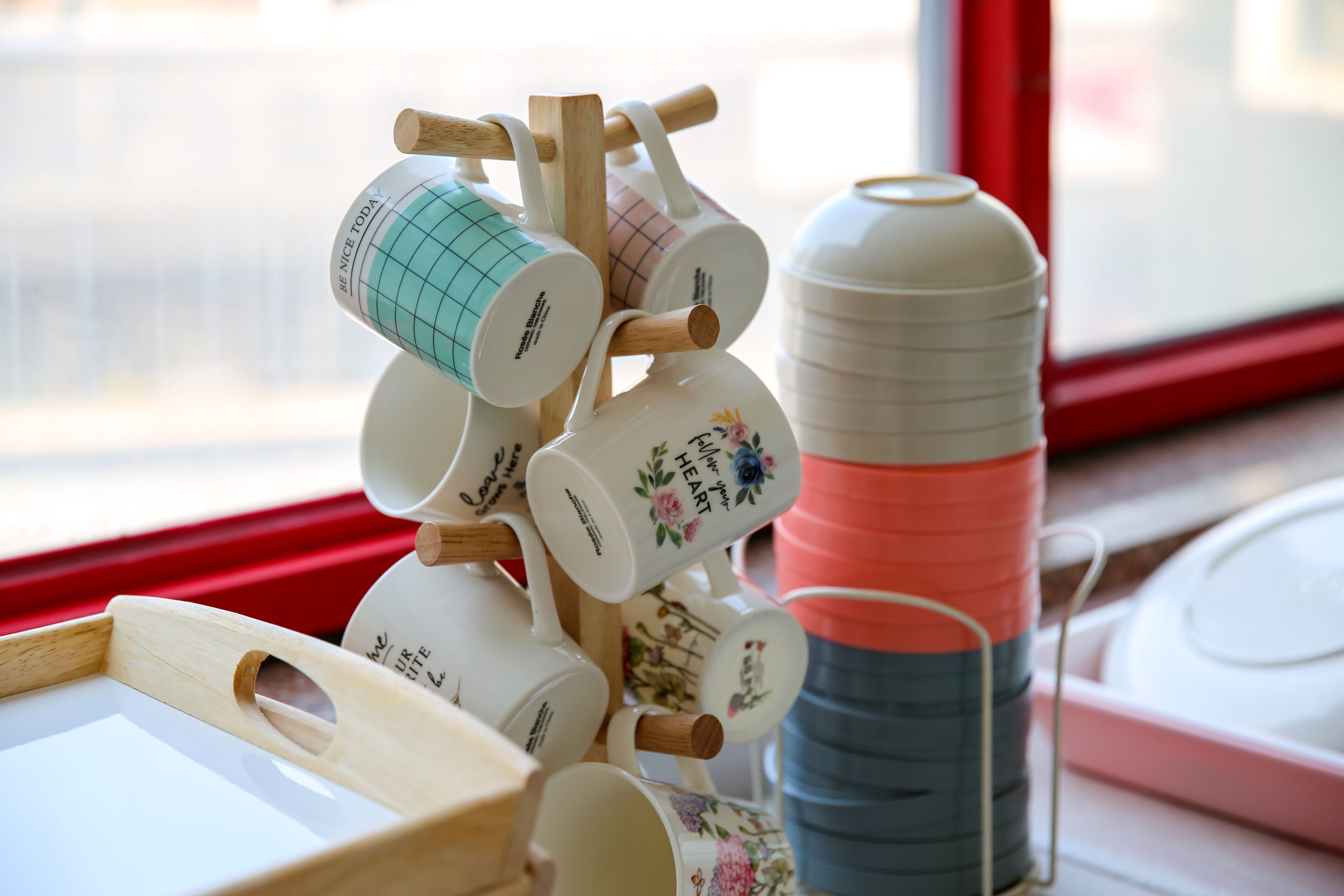 Una pila de tazas y un portavasos con diversos diseños y texto, sobre una mesa junto a una ventana.