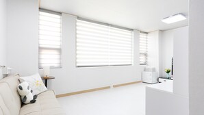 A minimalist room with a beige sofa, two large windows with blinds, a small bedside table with a lamp, and a white cabinet. - Dashim Guest House (Gunsan)