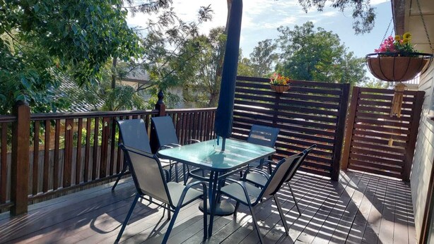 Eine überdachte Terrasse im Freien mit einem Glastisch und Metallstühlen, umgeben von einem Holzzaun und einer Holzterrasse.