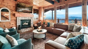 Ein gemütliches Blockhaus-Interieur mit Kamin aus Naturstein, Ledersofas und großen Fenstern mit Blick auf die Berge.