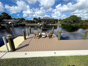 A wooden dock with chairs and a table, overlooking a calm body of water with a clear sky.