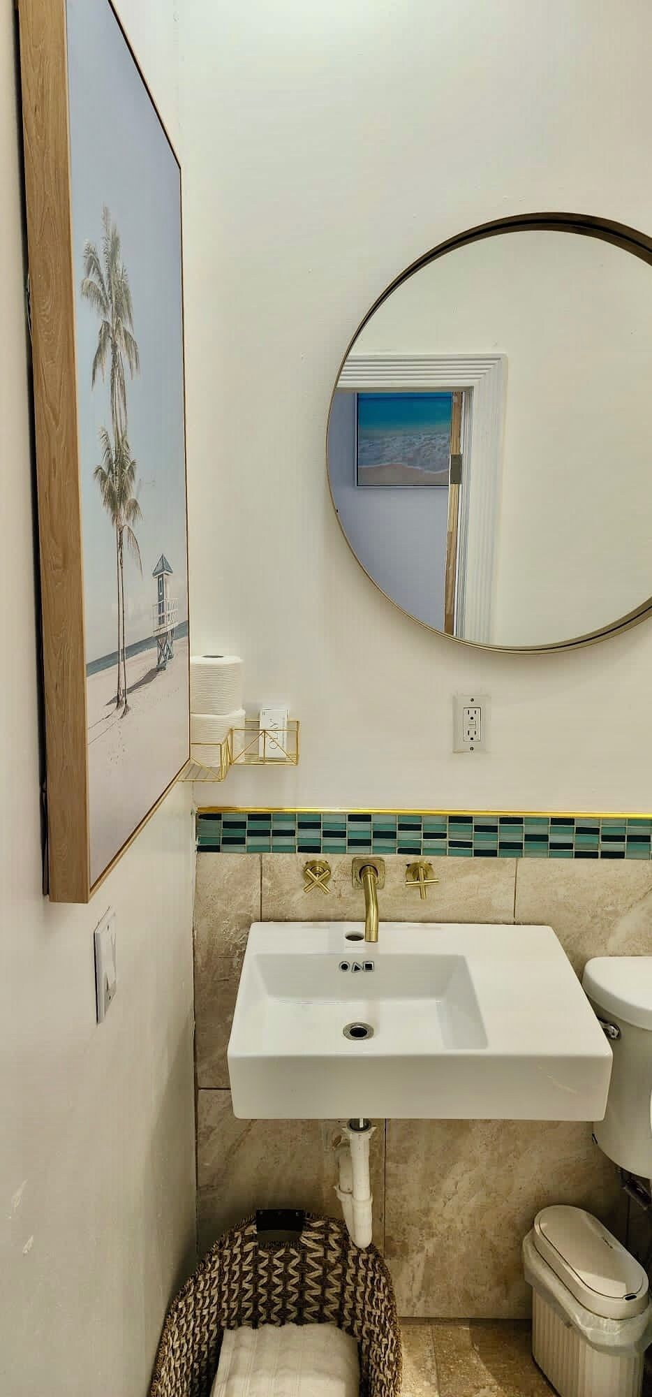 A bathroom with a white sink, a toilet, a trash bin, a towel rack, a framed picture, and a mirror.