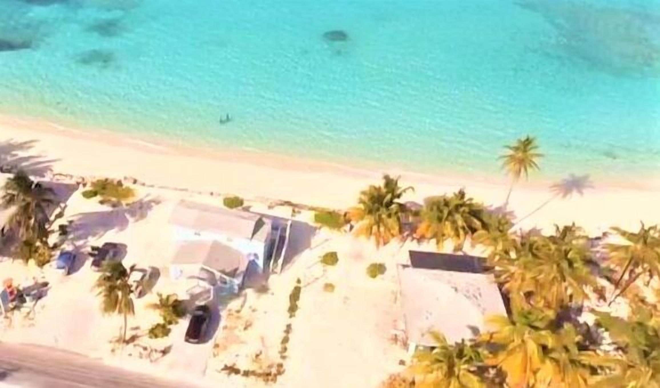 Una playa con agua turquesa cristalina, palmeras y algunos edificios.