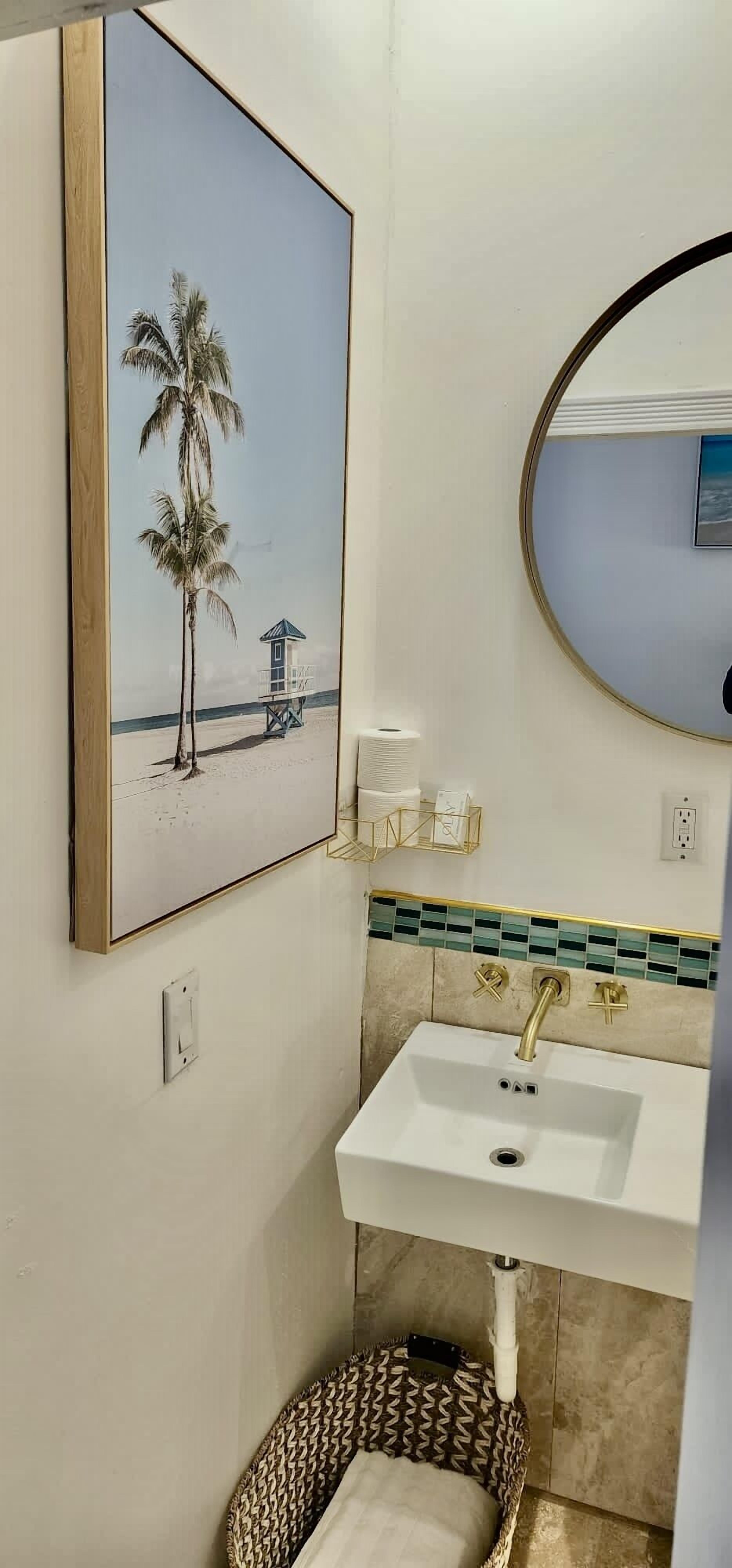 A bathroom with a beach-themed painting, a round mirror, a white sink with gold fixtures, and a patterned rug.
