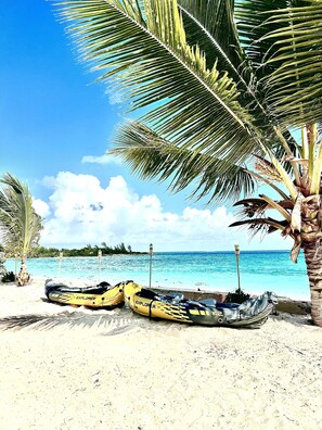 A beach with two yellow kayaks and a small boat on the sand, palm trees, and a clear blue sky.