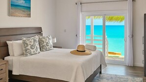 A bedroom with a bed, a nightstand, a wooden floor, a view of the ocean through a glass door, and a beach scene in the background. - The Suite Life (Rokers Point)