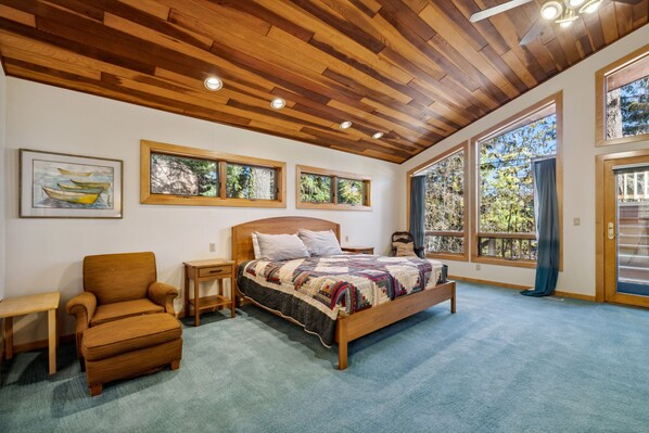 Un dormitorio con techo de madera, ventanas amplias, cama, silla y mesita de noche.