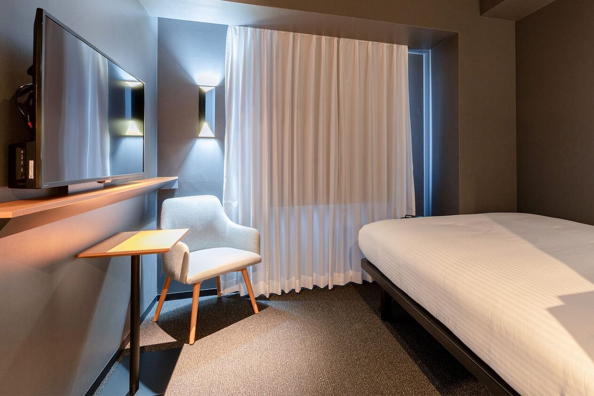 Quarto de hotel com uma cama, uma cadeira, uma televisão e uma janela com cortinas.