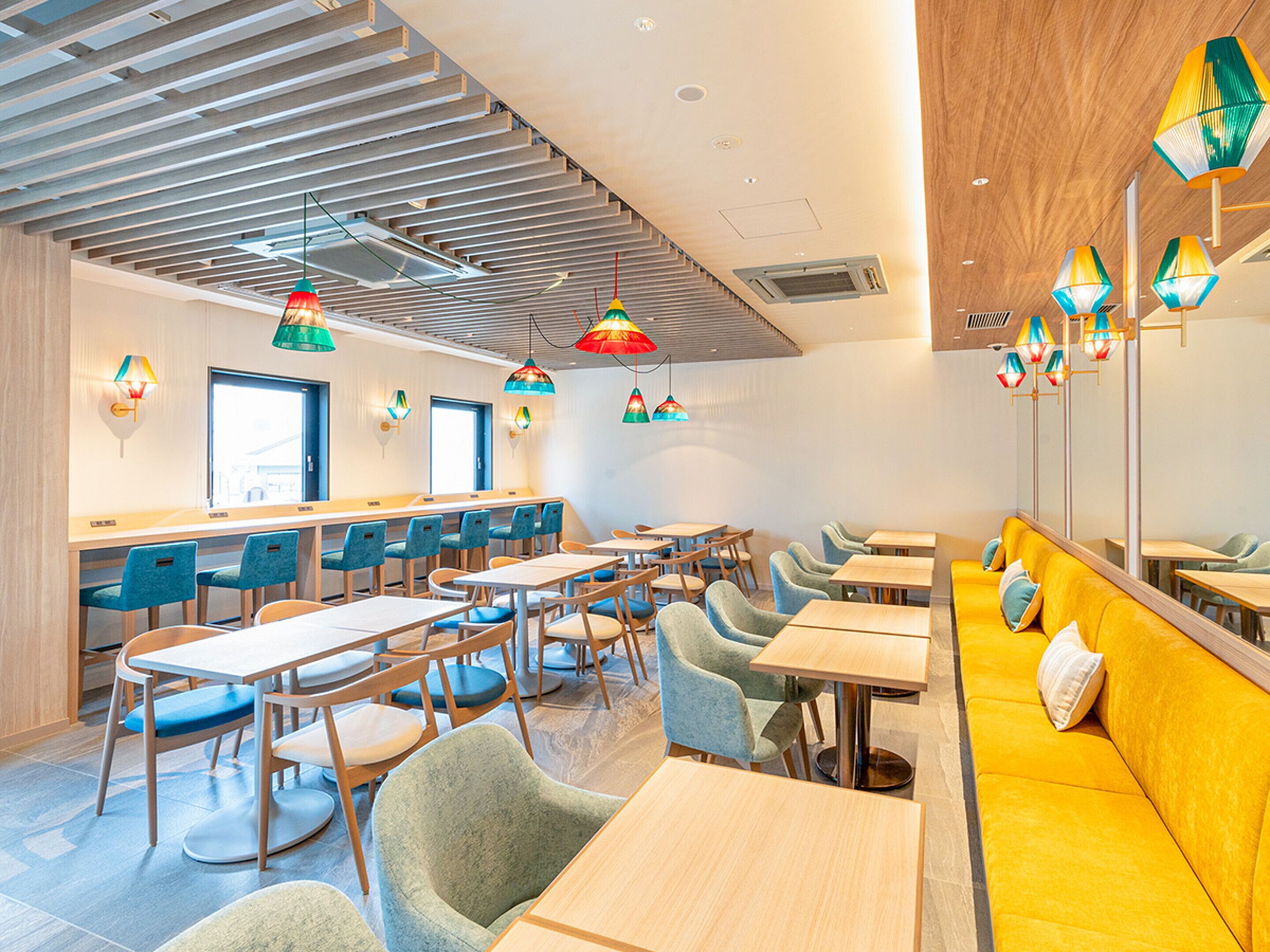A modern dining area with wooden tables and blue chairs, a yellow sofa, and colorful hanging lamps.