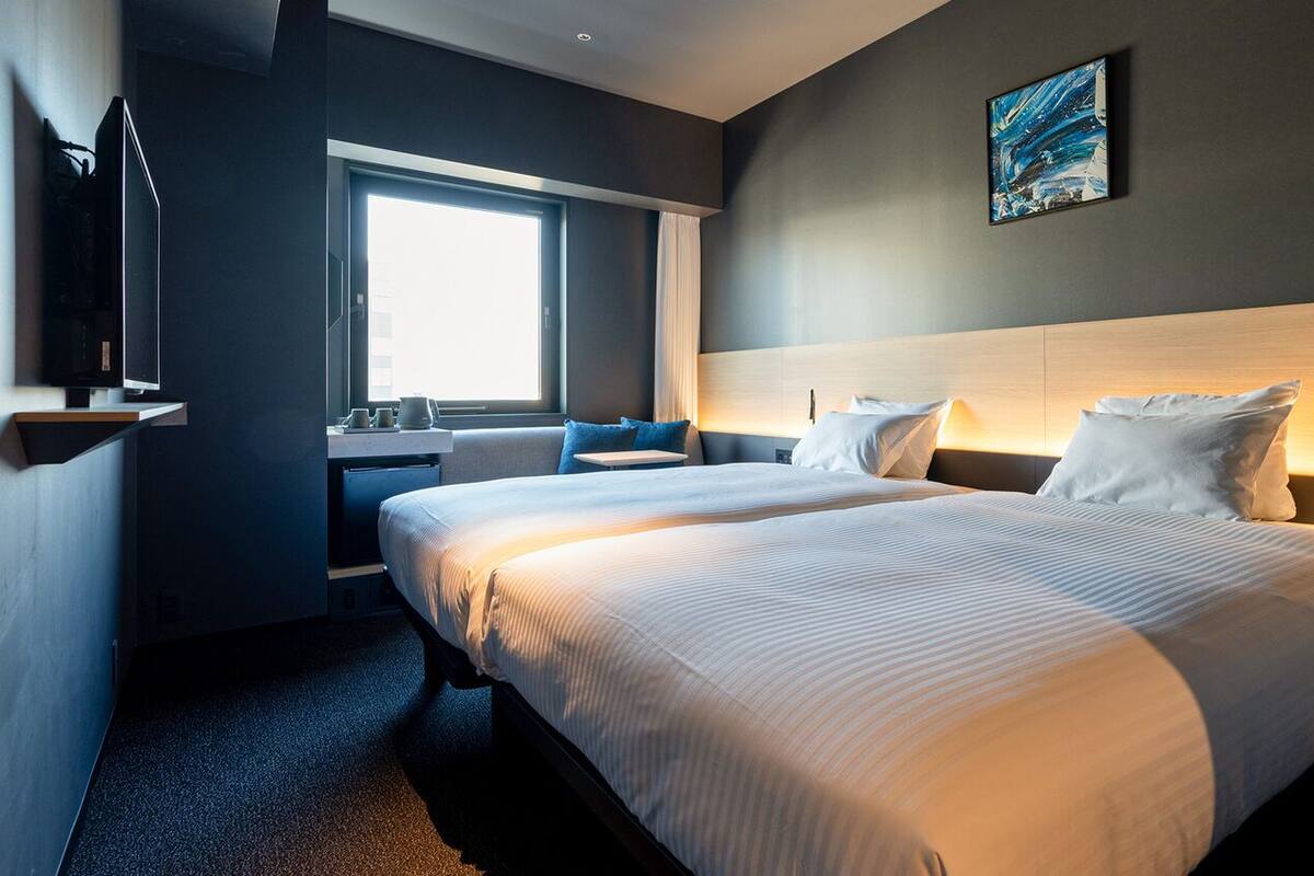 Quarto de hotel com duas camas, uma janela grande, um quadro na parede e uma escrivaninha com abajur.