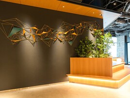Una moderna zona de recepción con una instalación artística geométrica en la pared, un macetero de madera con plantas y un mostrador de recepción bien iluminado.