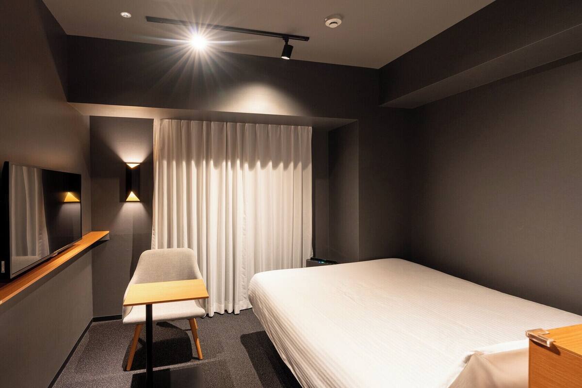 Quarto de hotel moderno com cama, escrivaninha, cadeira, televisão e janela com cortinas.