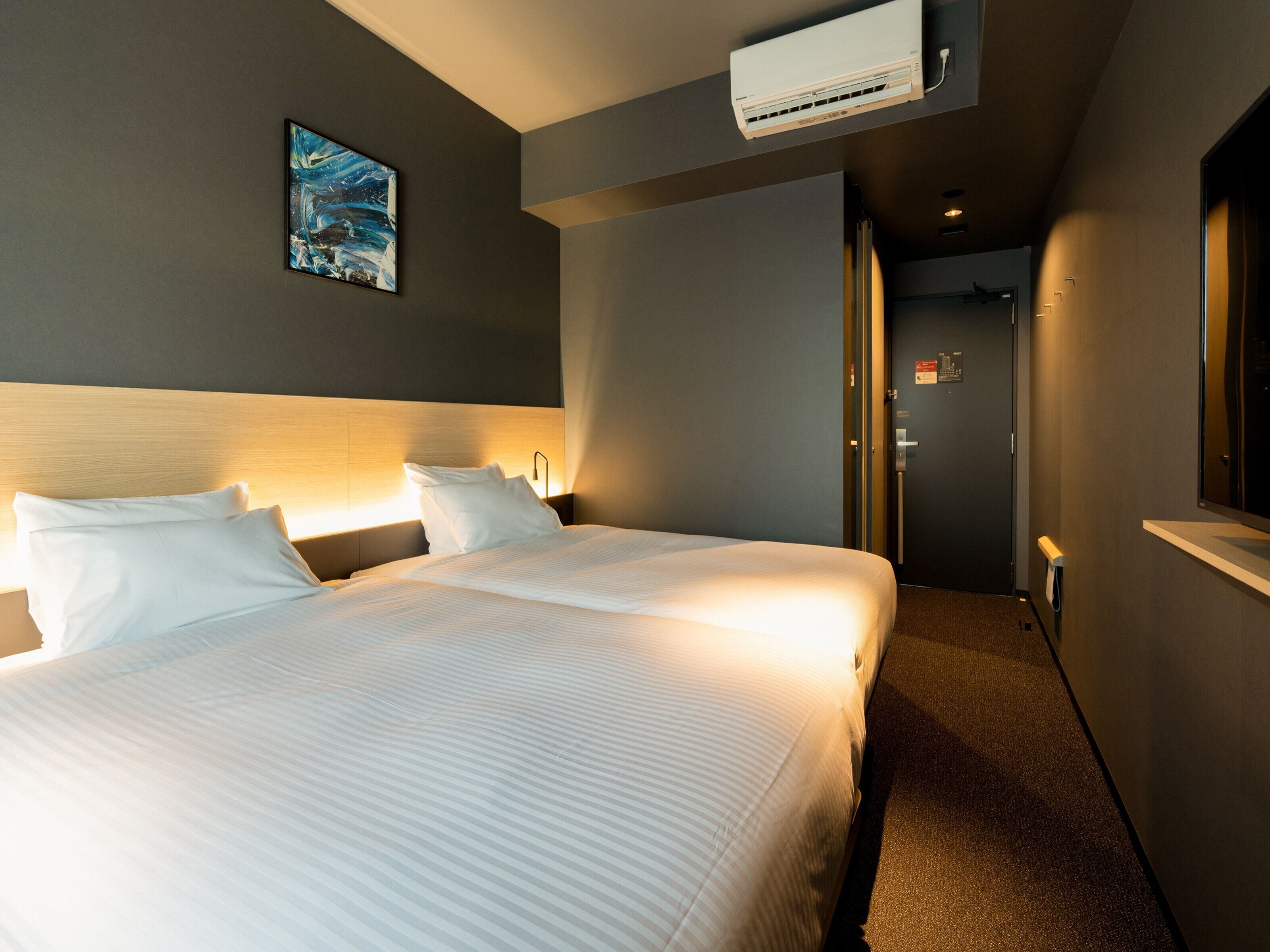 A hotel room with a large bed, a headboard, an air conditioning unit, a painting on the wall, and a door leading to another room.