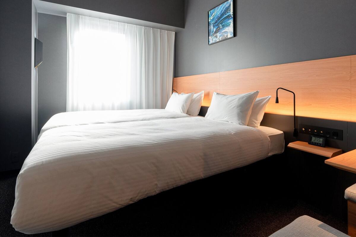 Quarto de hotel com uma cama grande, mesa de cabeceira com abajur, relógio e janela com cortinas.