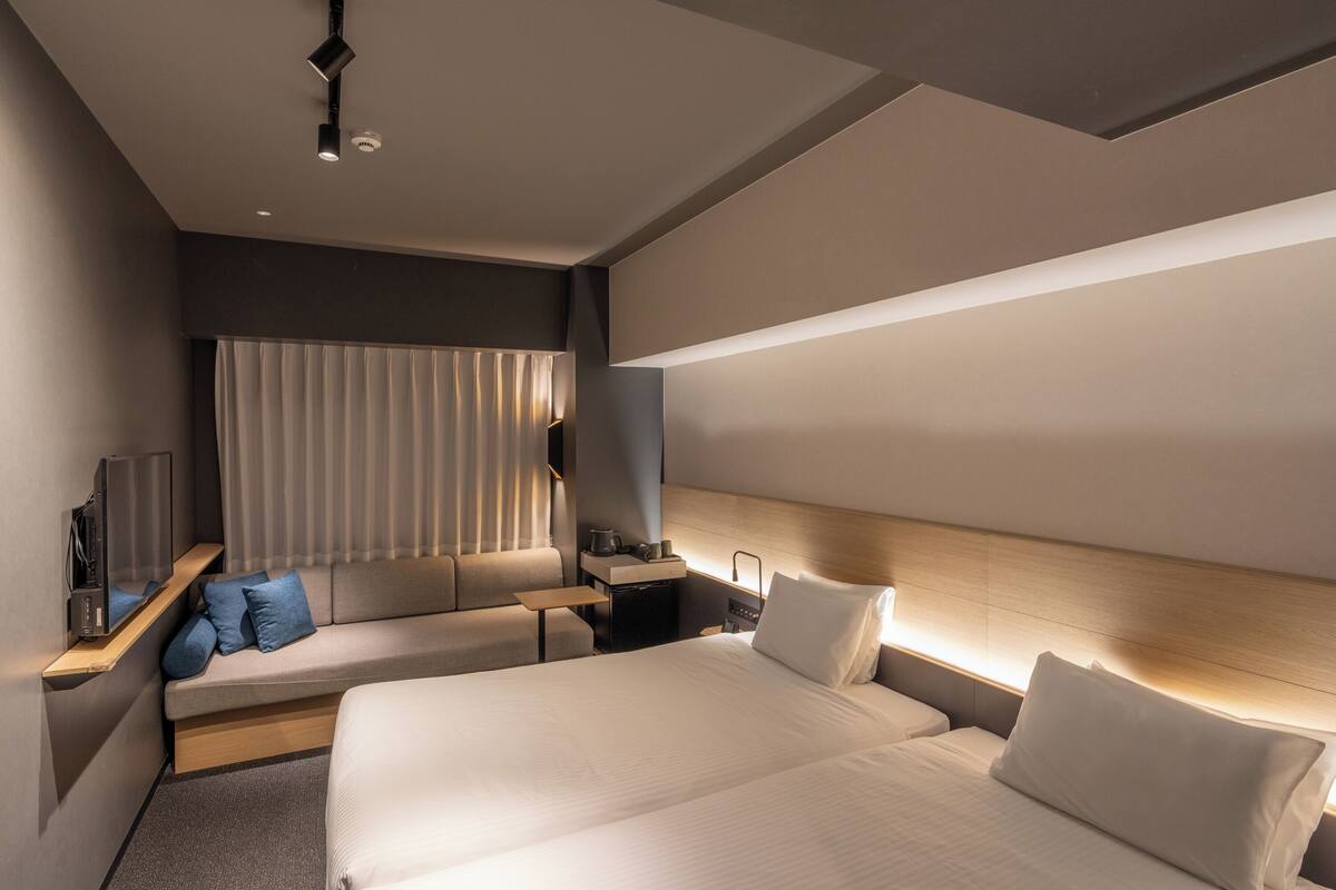 Quarto de hotel moderno com uma cama grande, área de estar com televisão e mesa de cabeceira com abajur.