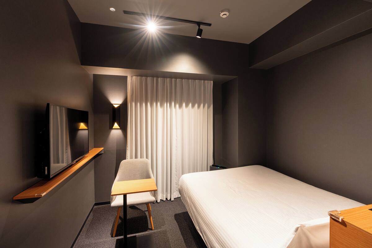 Quarto de hotel moderno com cama, escrivaninha, cadeira, televisão e uma janela grande com cortinas.