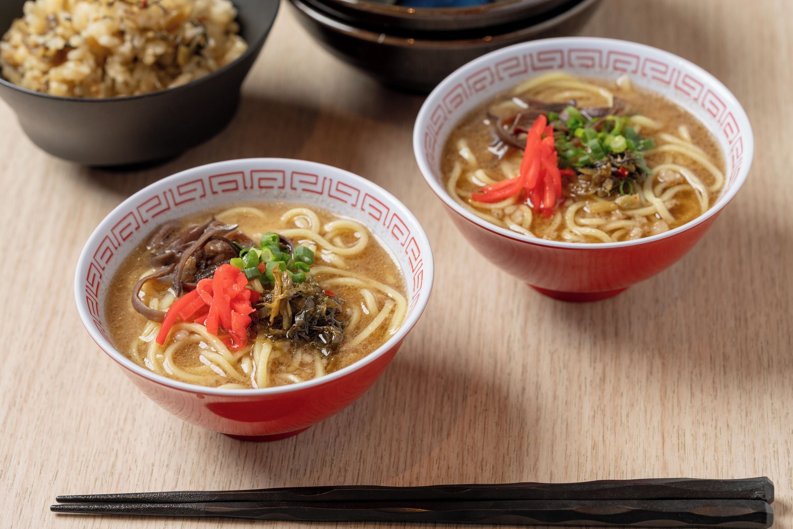 Two bowls of ramen with toppings, a pair of chopsticks, and a bowl of rice in the background.