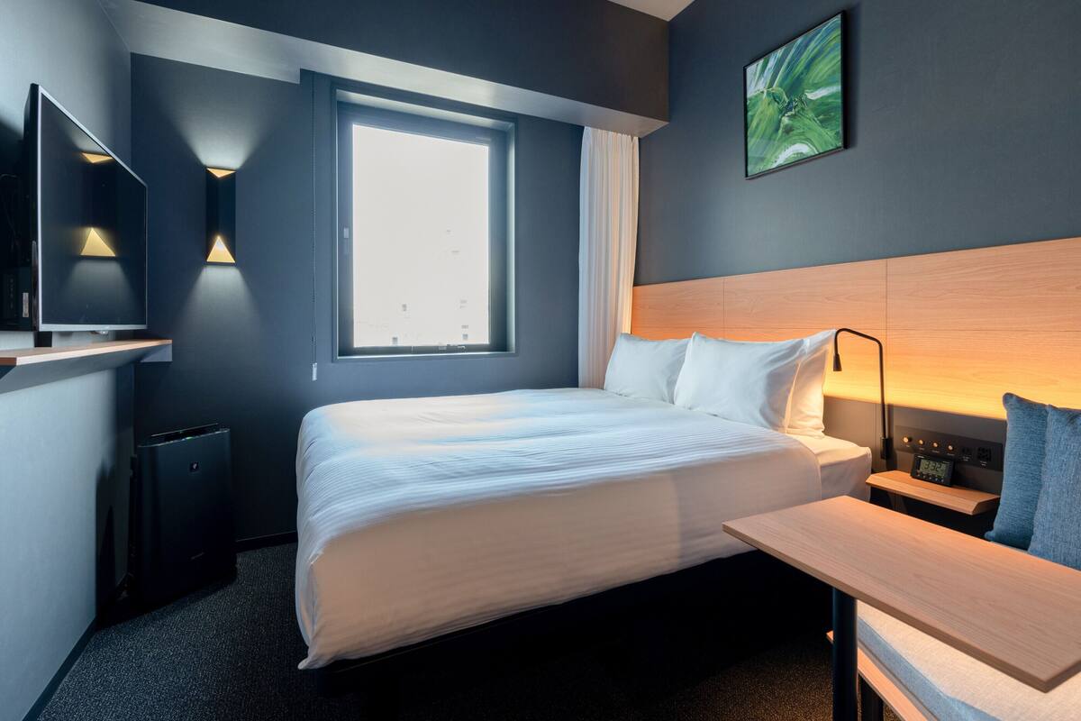 Quarto de hotel moderno com uma cama grande, uma escrivaninha, uma cadeira, televisão e janela com cortinas.