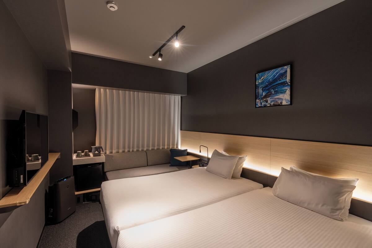 Quarto de hotel moderno com duas camas, uma escrivaninha, televisão e uma janela grande com persianas.