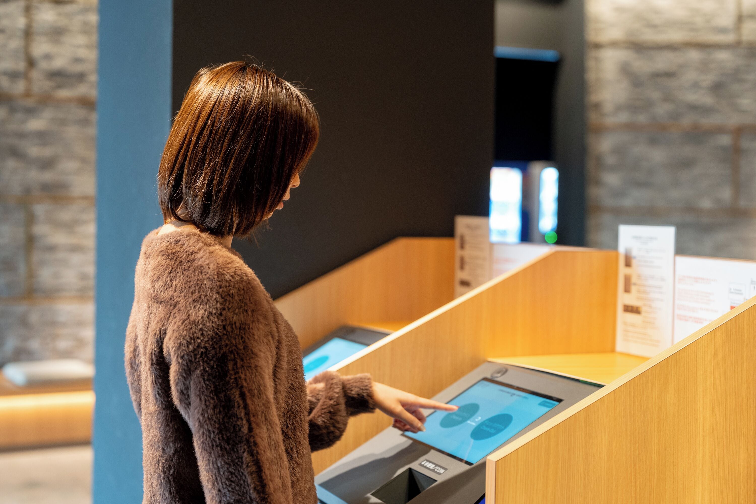 A person interacting with a touchscreen kiosk in a modern, well-lit room with stone walls and wooden furniture.