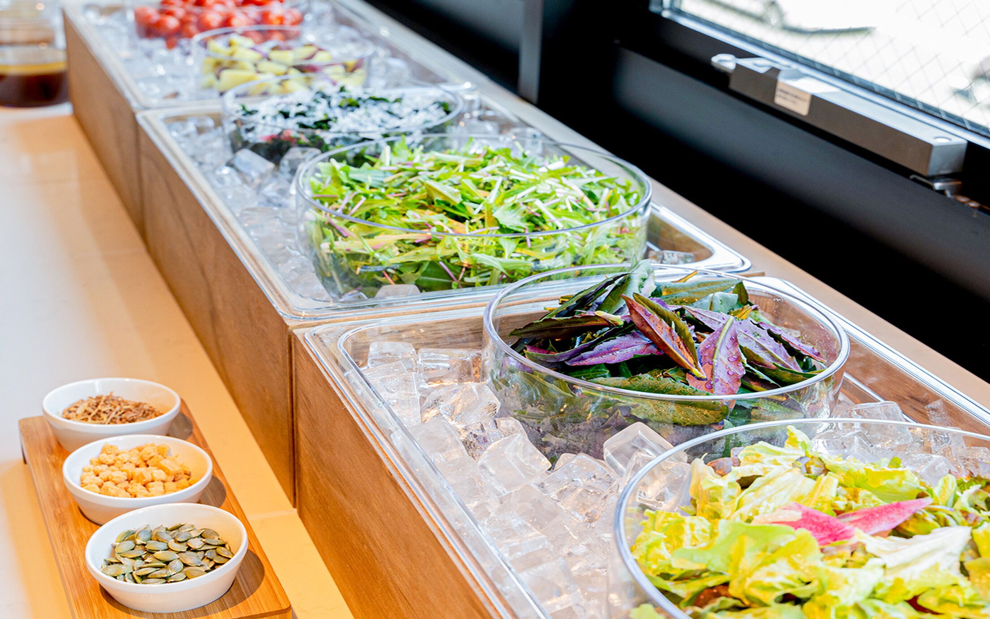 A buffet with various salads and toppings in clear containers, ice, and small bowls of seeds.