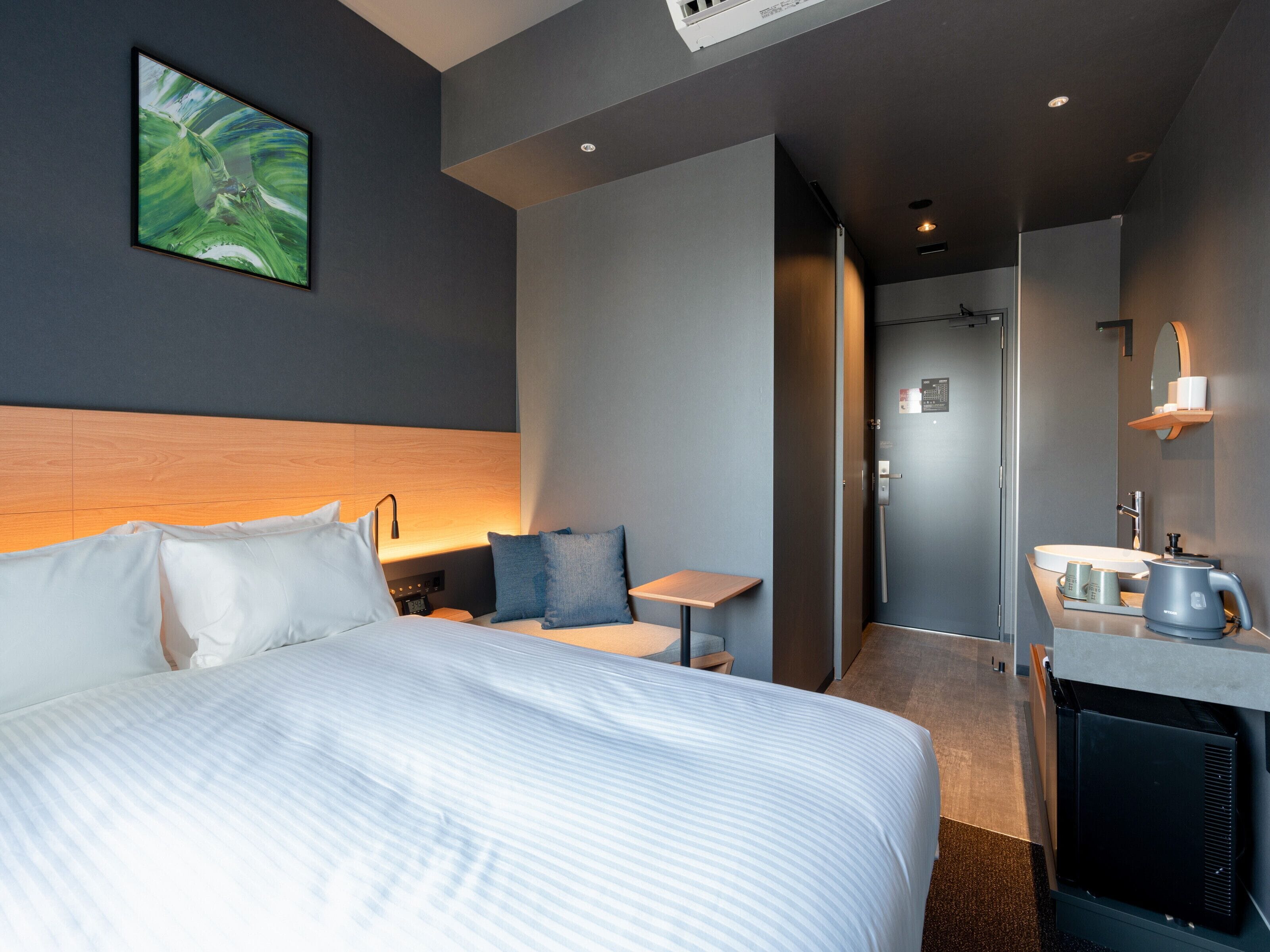 A modern hotel room with a large bed, a small desk, a seating area, and a bathroom with a mirror and sink.