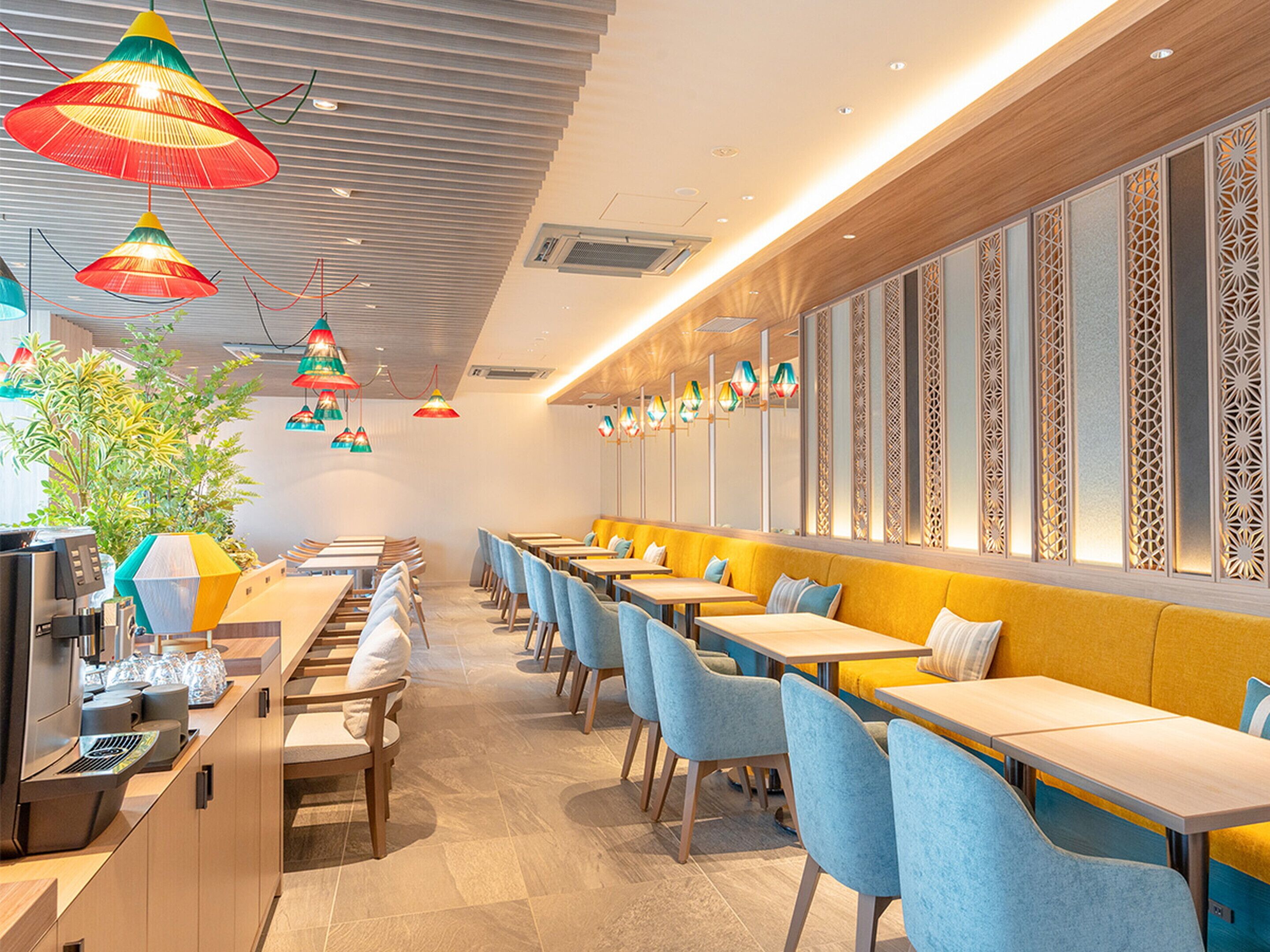 Een modern restaurantinterieur met lange gedeelde tafels, blauwe stoelen en kleurrijke hanglampen.