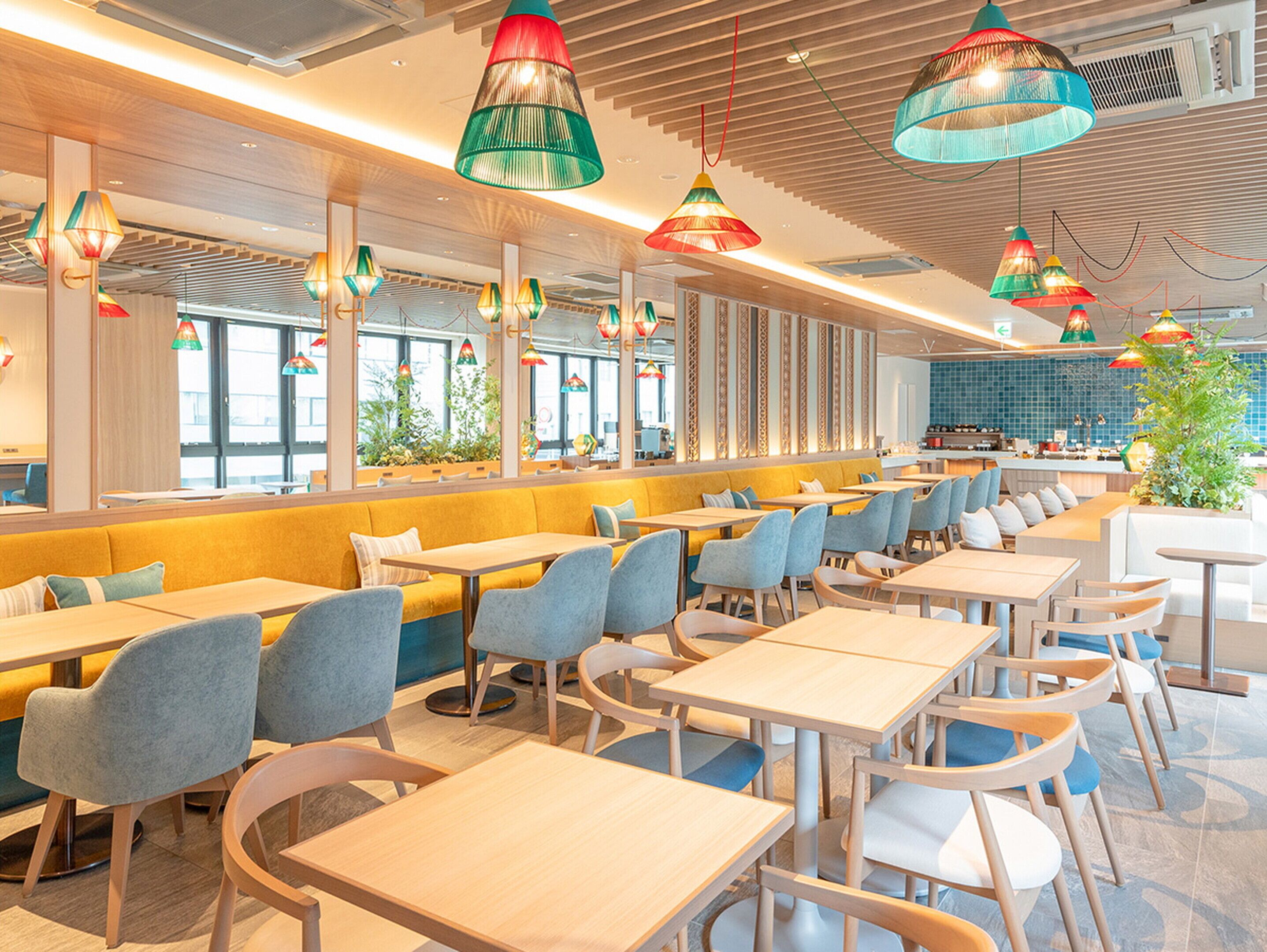 A modern restaurant with long communal tables and booths, colorful hanging pendant lights, and large windows.