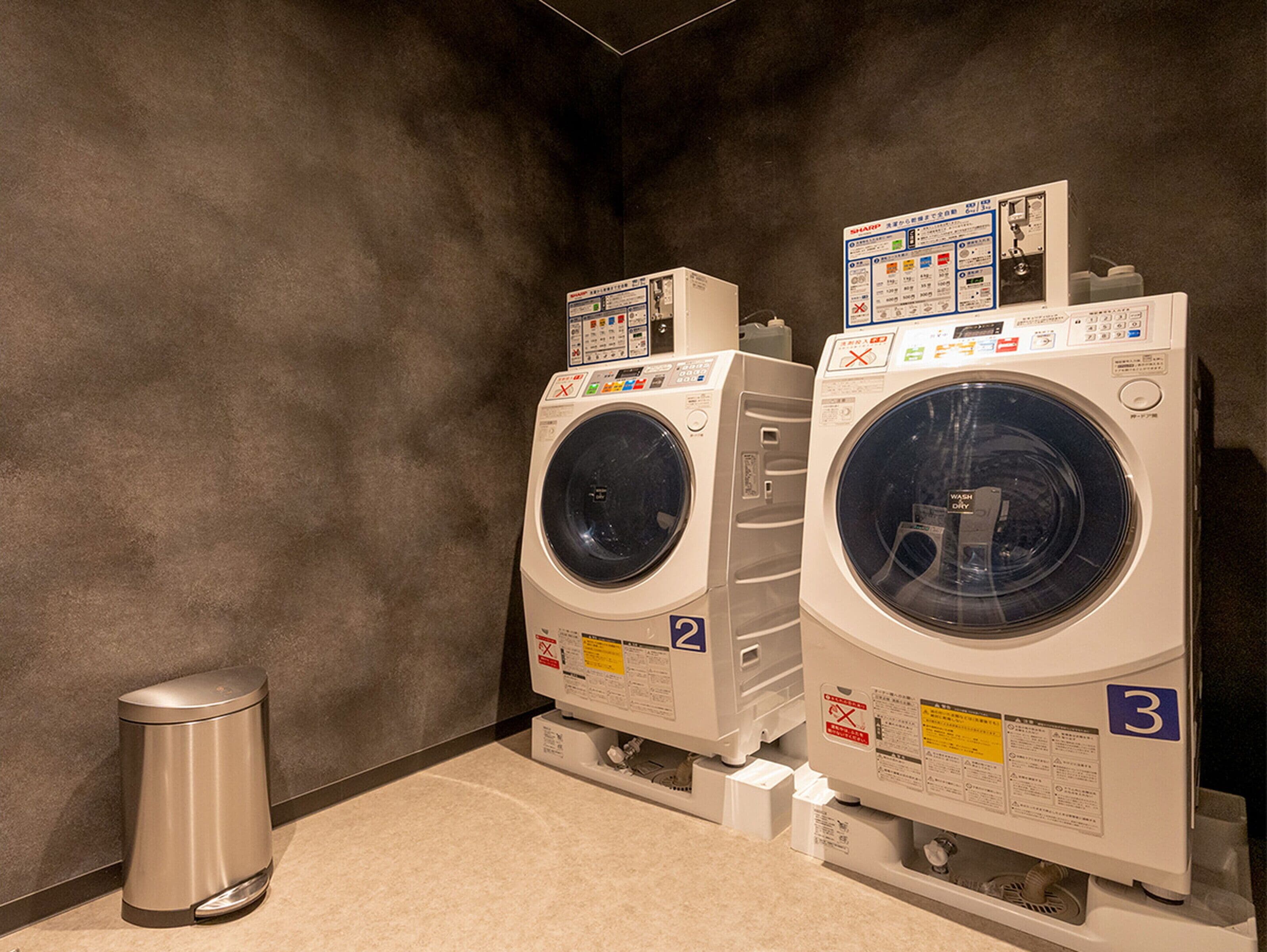 A laundry room with two front-loading washing machines, a trash can, and a wall-mounted control panel.