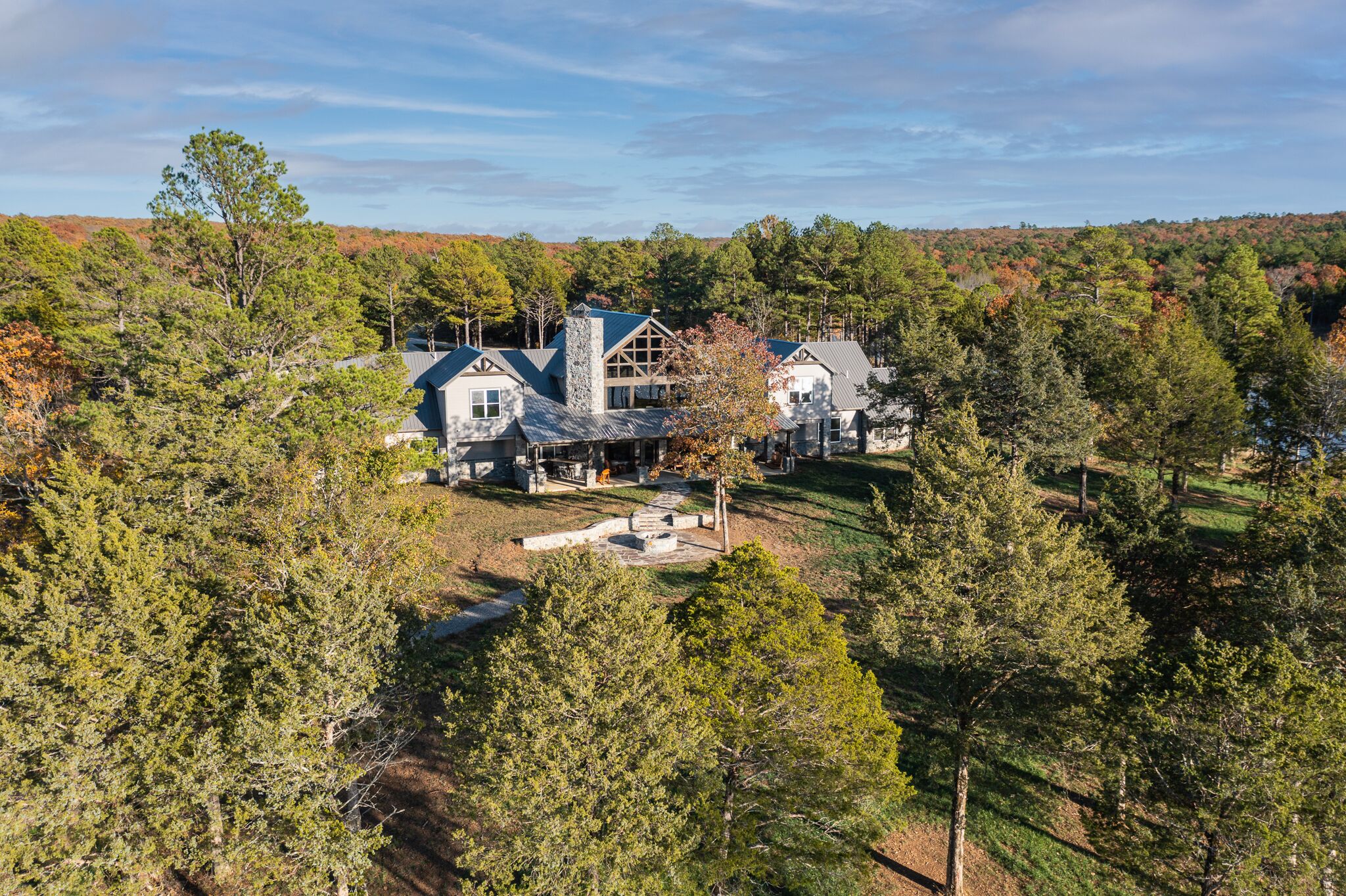 A large, two-story house with a stone chimney, surrounded by dense forest and a clear sky.